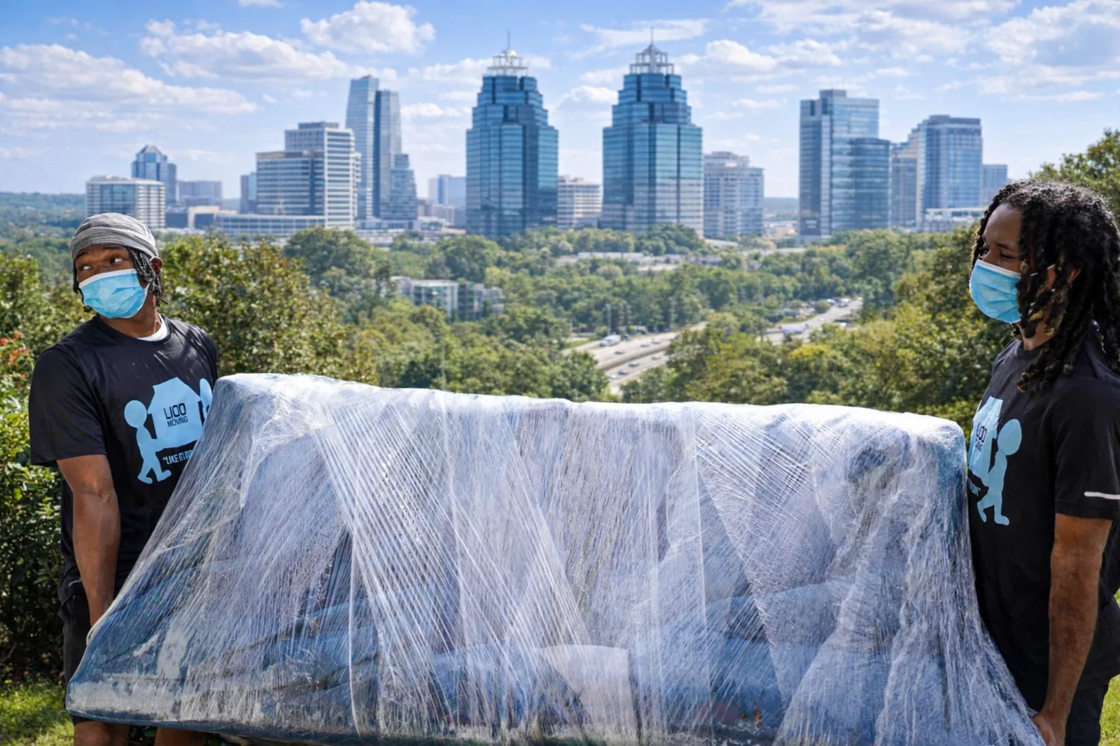 Two movers carry a shrink-wrapped sectional sofa with the Sandy Springs skyline behind them