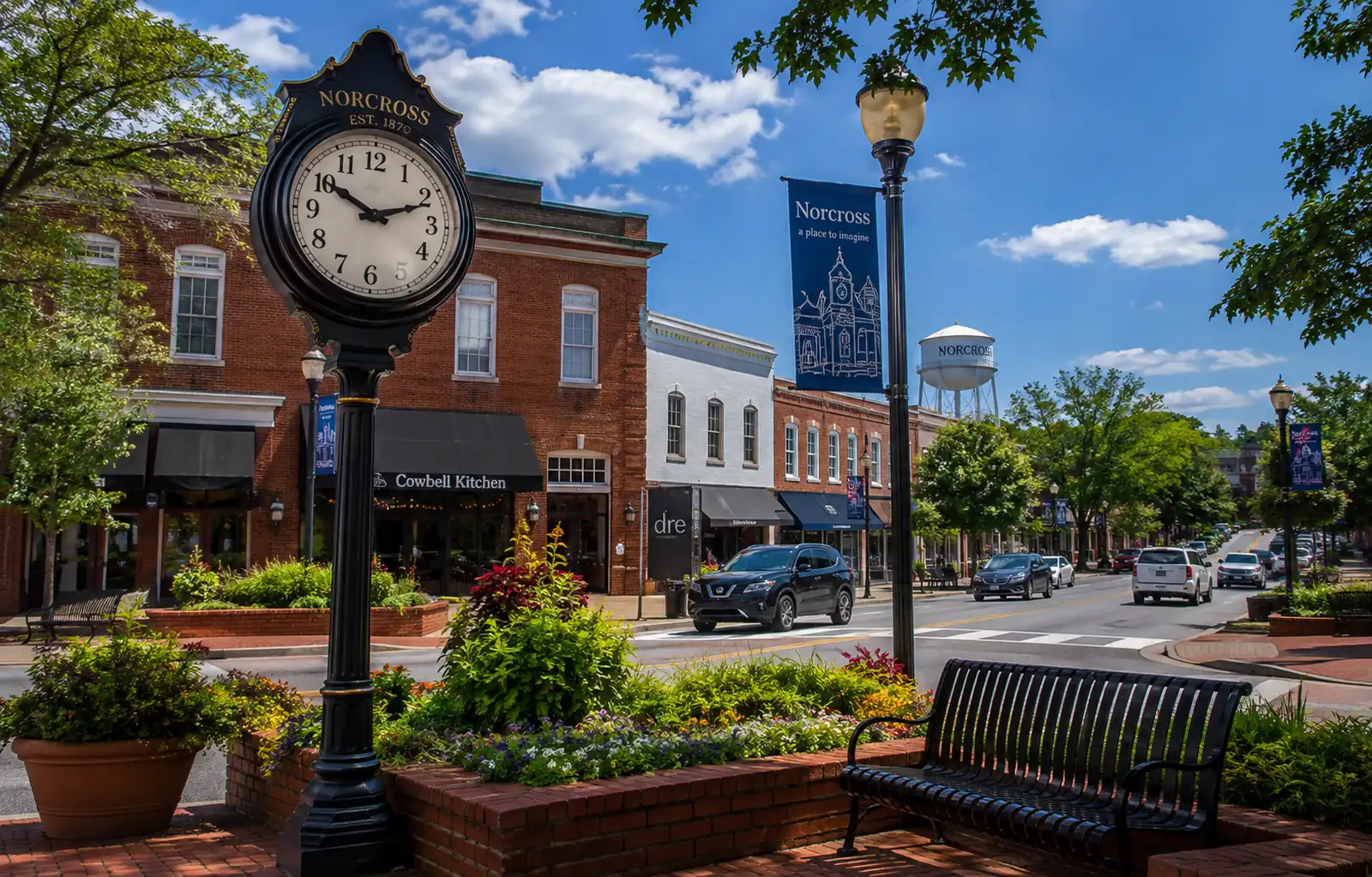 Norcross clock stands beside landscaped planters with brick storefronts and a downtown roadway