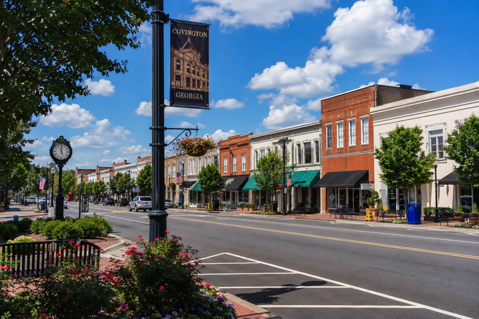 Historic brick storefronts, landscaped sidewalks, and a downtown street in Covington, Georgia beneath a bright blue sky.