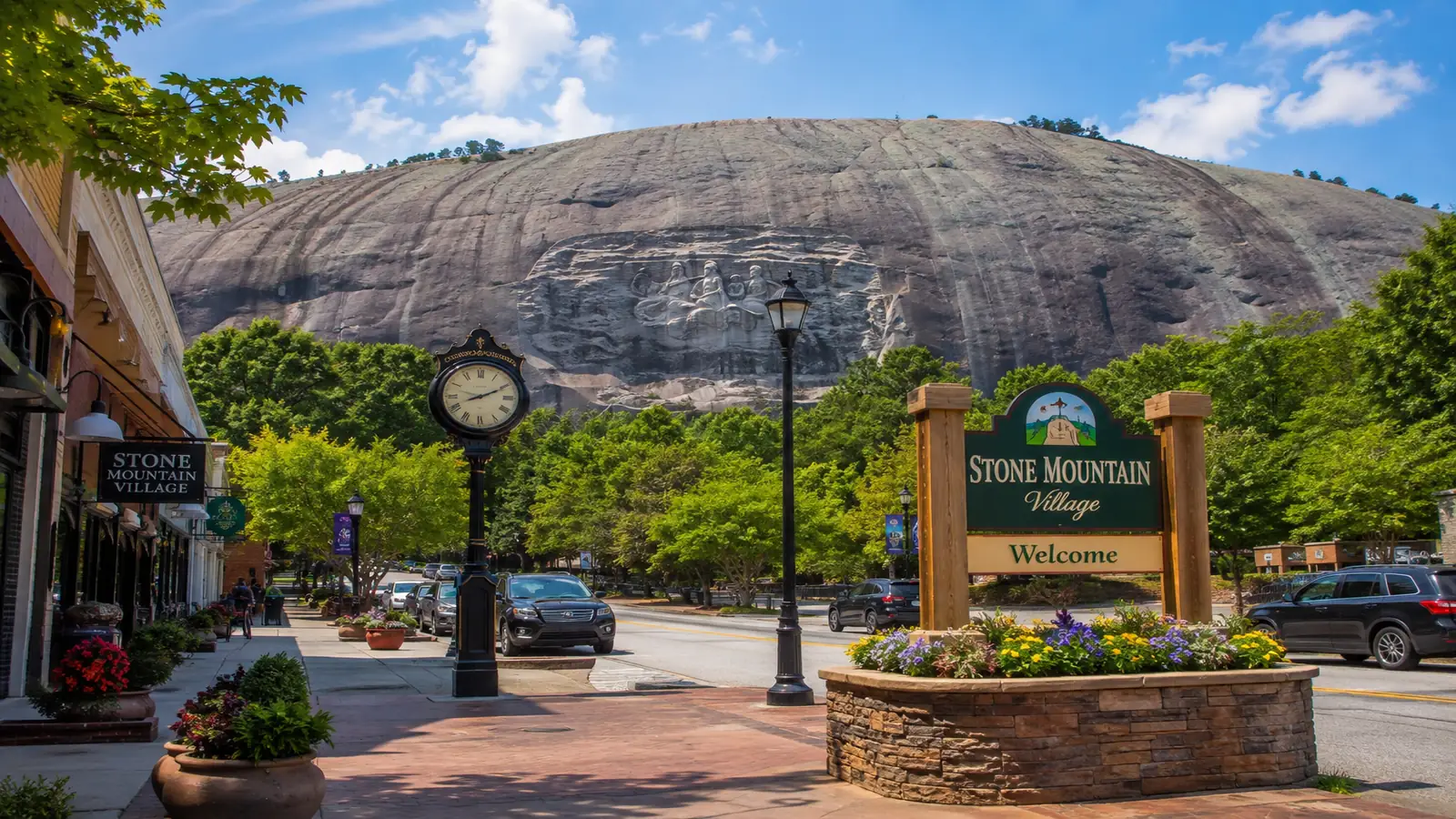 Stone Mountain Village streetscape with brick storefronts, cars, trees, and the granite mountain beyond.