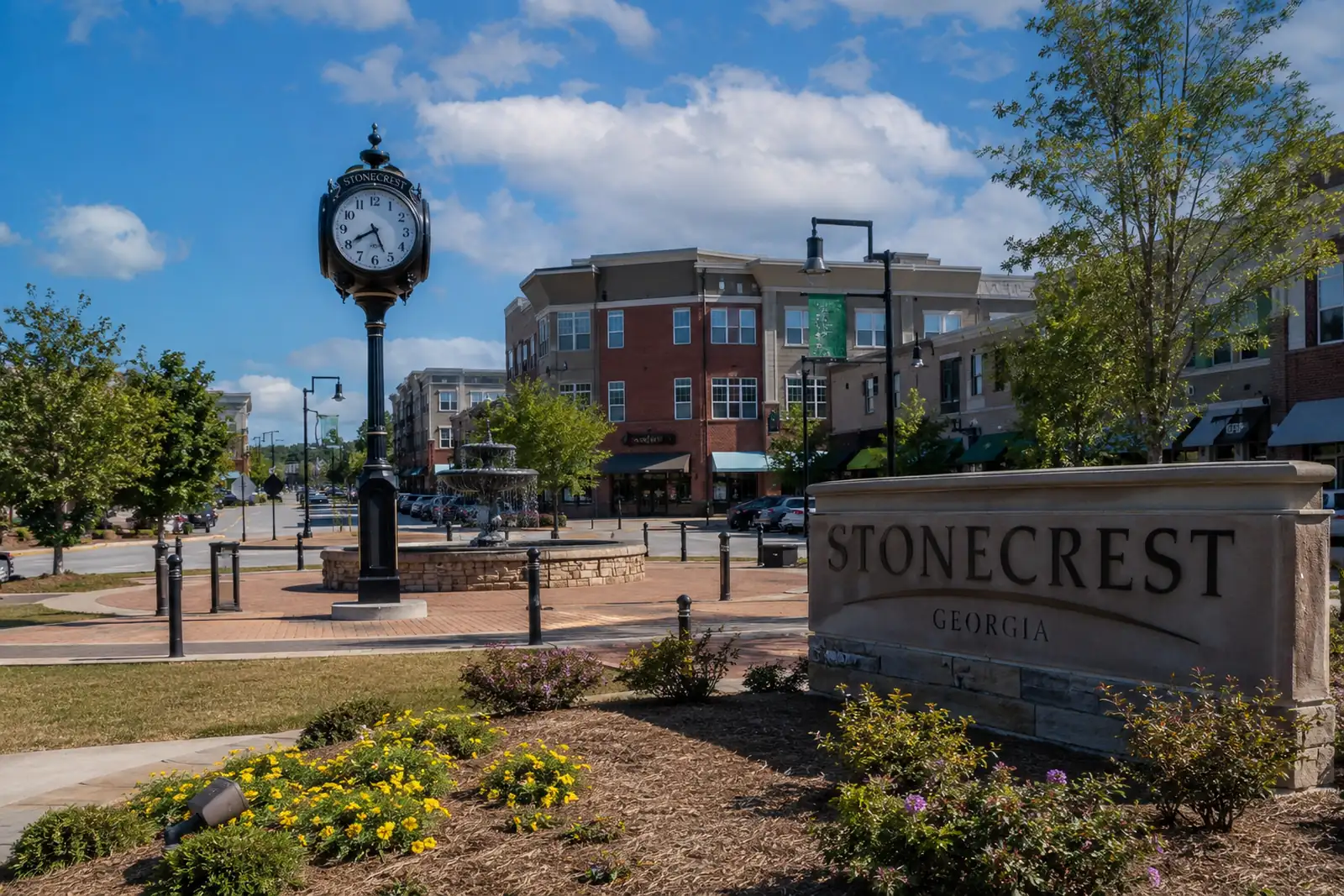 Stonecrest Georgia monument beside a landscaped plaza, fountain, clock, sidewalks, and mixed-use buildings
